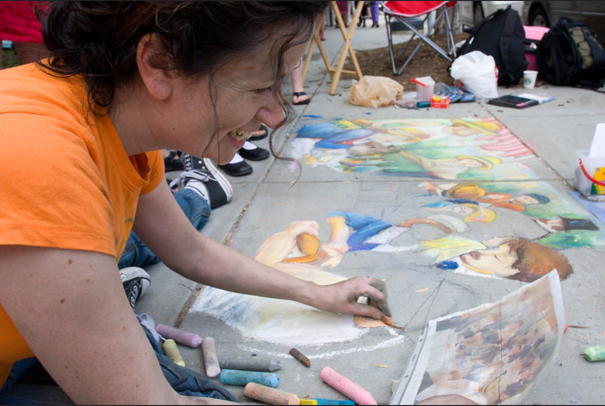 Dana Ellyn laughing while working on the chalk recreation, onlookers and folding chairs visible behind her