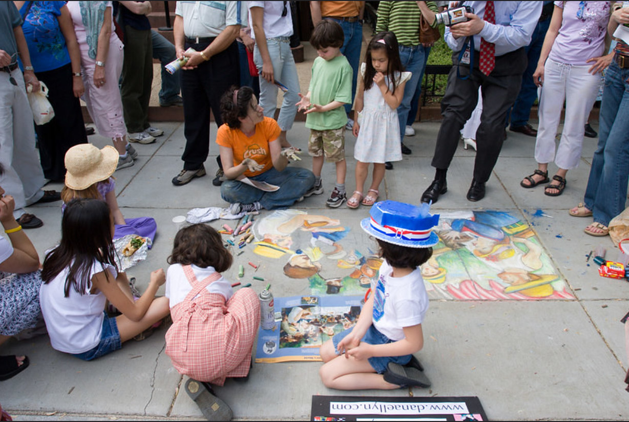 Children sit beside the finished sections of the chalk recreation while Dana Ellyn talks with more kids and a bystander films with a video camera, a sign reading www.danaellyn.com at the edge of the piece